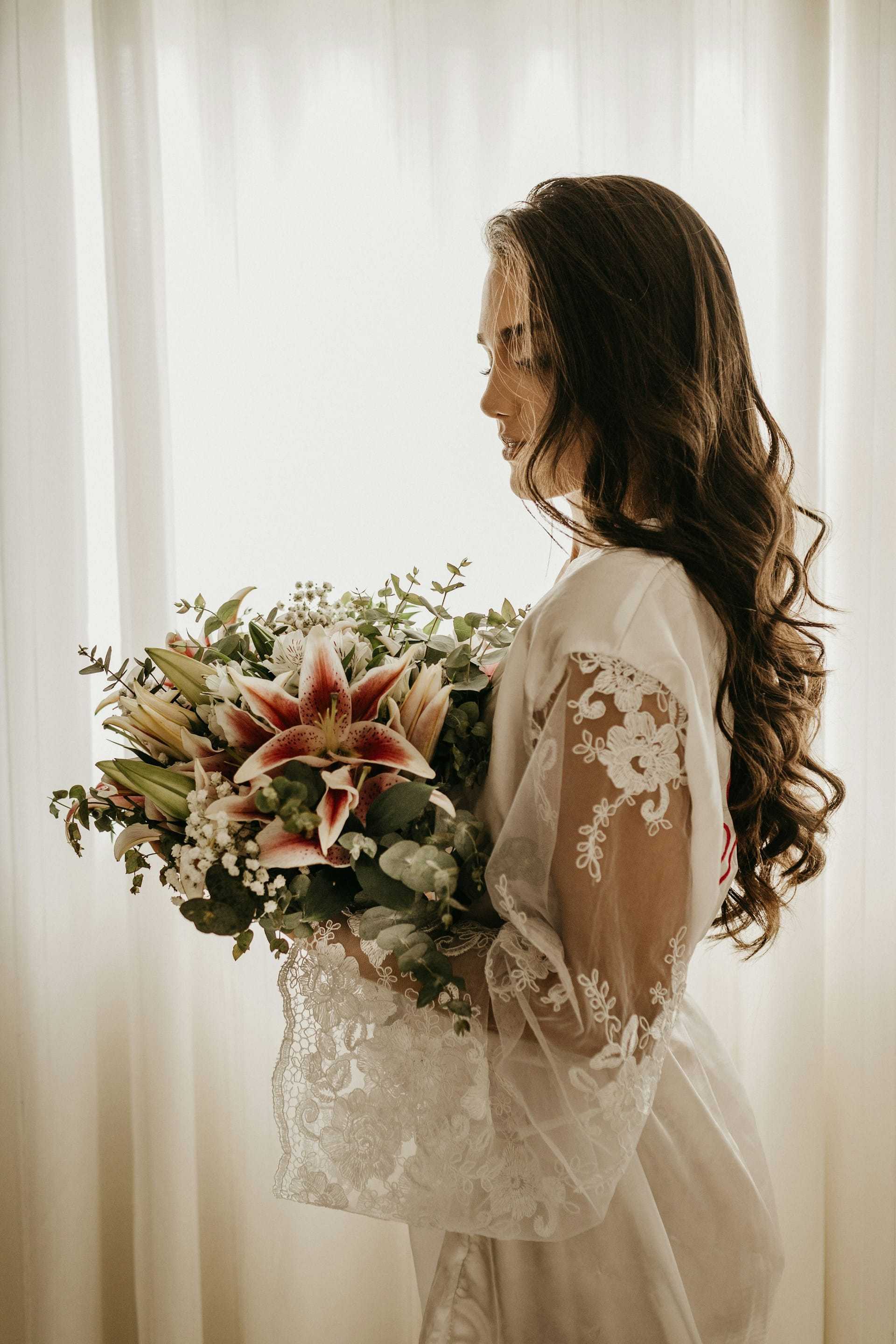 Bride in lace robe holding a large floral bouquet, standing by a softly lit window.